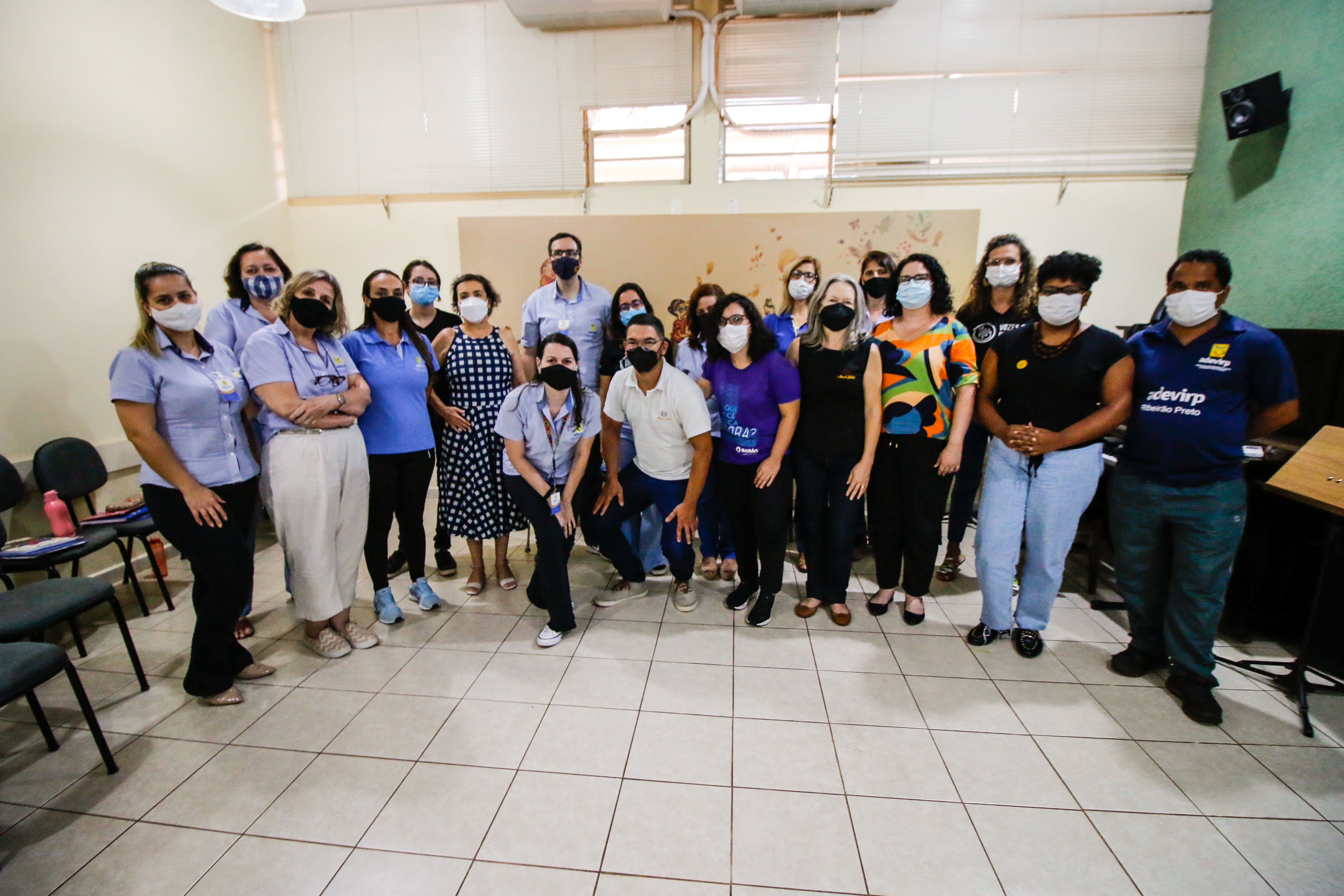 Fotografia em plano geral, apresenta os participantes do treinamento em uma sala ampla com paredes e chão claros. 
Todos estão em pé, um ao lado do outro, usam máscaras e olham para frente.