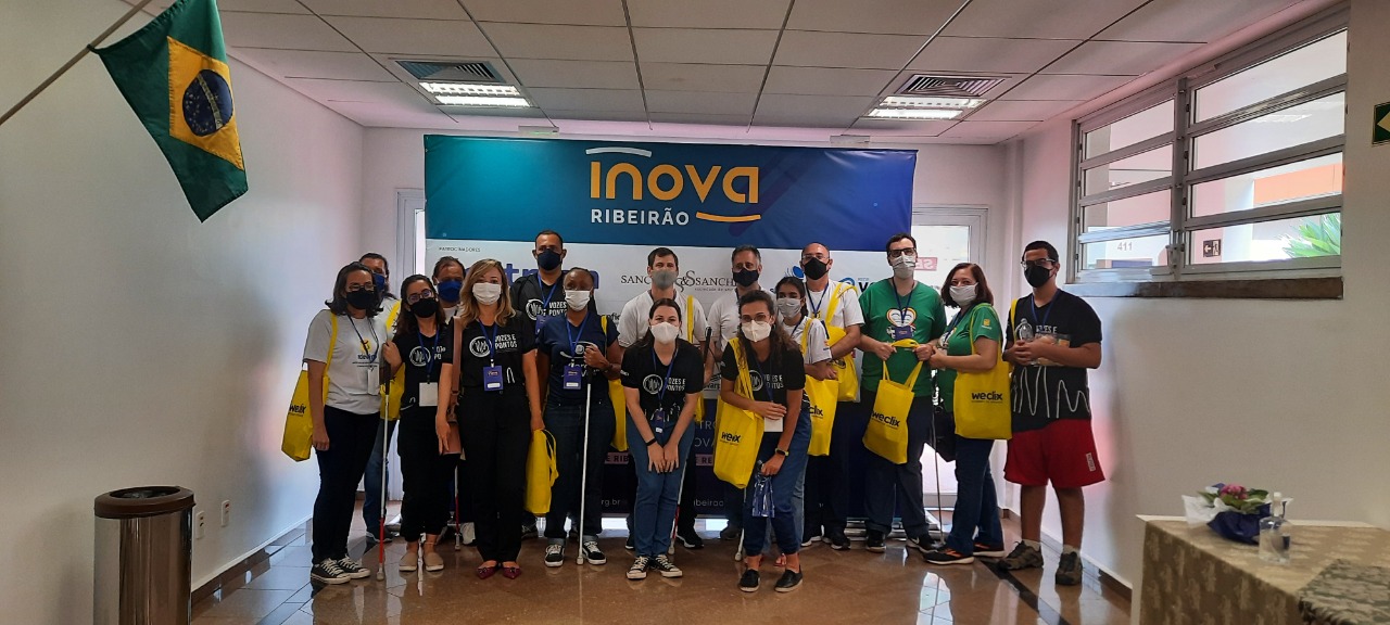 Fotografia colorida, em plano geral, mostra os educandos e alguns profissionais da Adevirp em frente ao banner do Inova Ribeirão, em um ambiente fechado com paredes brancas. Todos estão em pé, bem próximos um do outro, usam máscara, olham para a foto e sorriem. Alguns usam o uniforme preto do projeto Vozes e Pontos e outros usam o uniforme branco ou verde da Adevirpi.