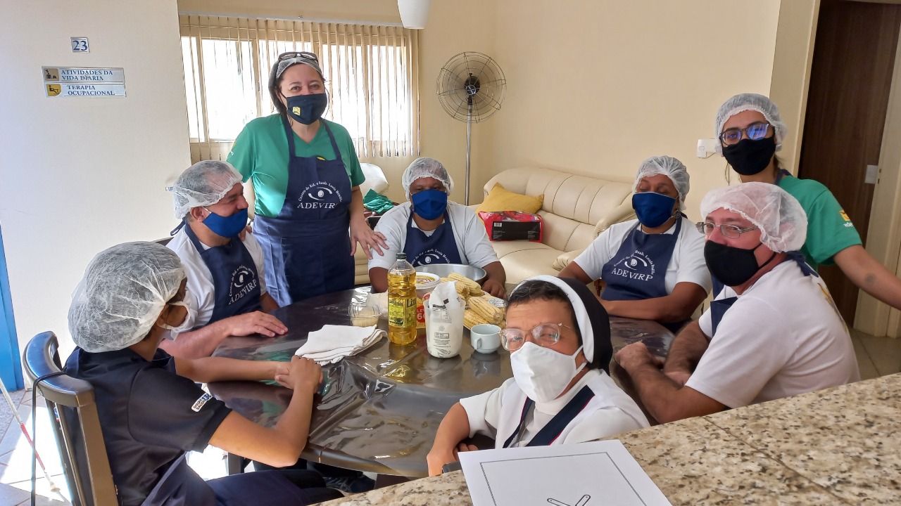 Fotografia colorida mostra cinco educandos e três profissionais da Adevirpi, dentro da sala de Atividades da Vida Diária AVD, em volta de uma mesa de madeira redonda.
Ao fundo, paredes claras, uma janela com persiana, um ventilador e um sofá da cor bege claro. Sobre a mesa estão os ingredientes e utensílios: óleo, milho, leite, queijo, uma forma redonda e uma xícara branca de chá.
Todos usam máscara, touca descartável e olham para a foto e sorriem.